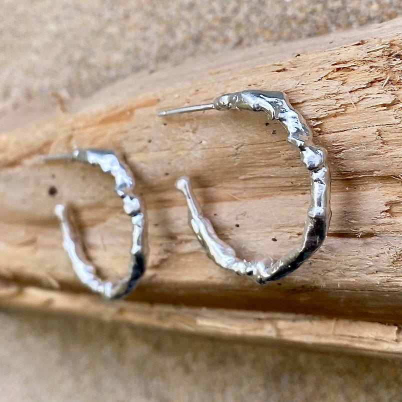 Silver statement hoop earrings gleam on a piece of driftwood on a Cornish beach, adorned with intricate textures crafted by the distinct process of seawater casting. Inspired by the sea and intricately formed within it, these earrings captivate with their undulating surfaces that gracefully reflect the light with every movement.