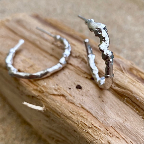 Silver statement hoop earrings gleam on a piece of driftwood on a Cornish beach, adorned with intricate textures crafted by the distinct process of seawater casting. Inspired by the sea and intricately formed within it, these earrings captivate with their undulating surfaces that gracefully reflect the light with every movement.