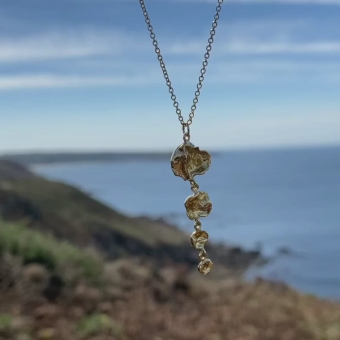 A golden necklace dances in the breeze along the Cornwall coastline, framed by the vast sea in the background. Its brilliant glow is accentuated by the sunlight, showcasing its distinctive shape and texture. This necklace, suspended on a chain, draws inspiration from the sea and nature, crafted in Cornish seawater.