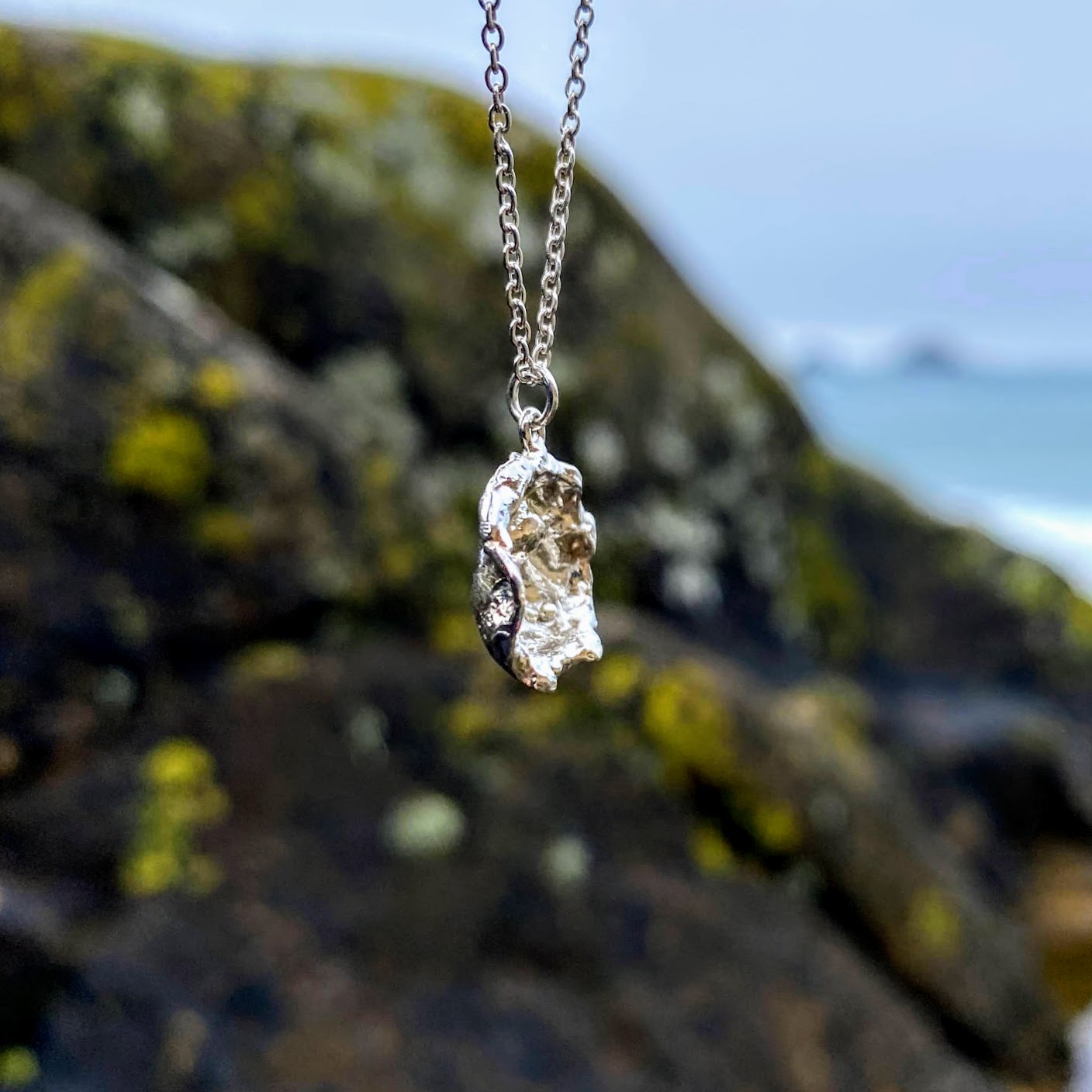 Imagine a stunning sterling silver pendant inspired by the sea, featuring a natural texture and charming organic shape. Crafted by pouring molten silver into the sea, its unique form captures the essence of ocean waves. As sunlight dances upon it, the pendant comes alive, showcasing its intricate details and textures. Picture it photographed on a picturesque beach in Cornwall, close to where it was lovingly created.