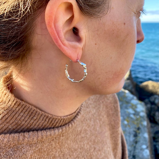 Silver statement hoop earrings gleam along the coastal shores of Cornwall, adorned with intricate textures crafted by the distinct process of seawater casting. Inspired by the sea and intricately formed within it, these earrings captivate with their undulating surfaces that gracefully reflect the light with every movement.