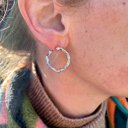 Shiny sterling silver hoop earrings being worn. The unique hoop is worn flat against the earlobe and twists around so that the end is behind the ear. Sparkling in the sunlight highlighting the natural and heavily textured form, achieved by casting in to Cornish seawater. Handmade in Cornwall by jewellery designer Joseph Lamsin from Penzance, who is inspired by the sea and his natural surroundings, and makes jewellery using his natural surroundings for his sea themed collections.